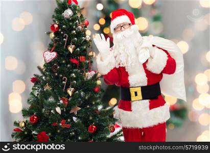 christmas, holidays and people concept - man in costume of santa claus with bag and christmas tree waving hand over lights background