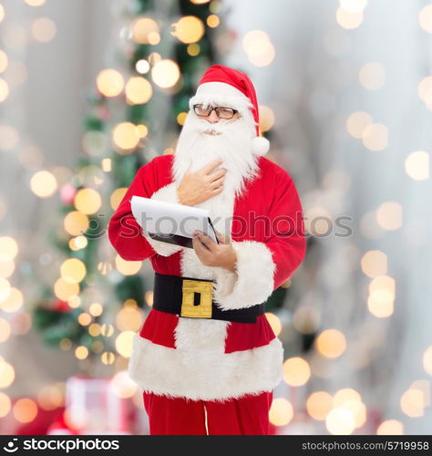 christmas, holidays and people concept - man in costume of santa claus with notepad over tree lights background