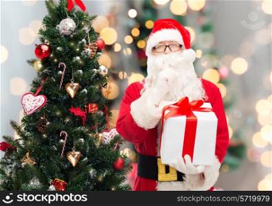 christmas, holidays and people concept - man in costume of santa claus with gift box and tree making hush gesture over lights background