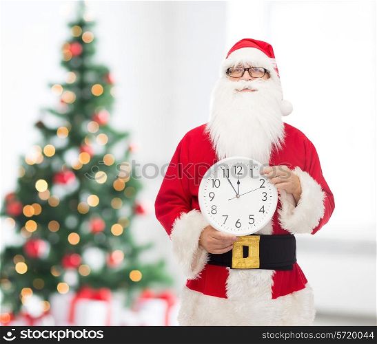 christmas, holidays and people concept - man in costume of santa claus with clock showing twelve pointing finger over living room with tree background