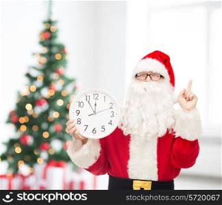 christmas, holidays and people concept - man in costume of santa claus with clock showing twelve pointing finger up over living room with tree background