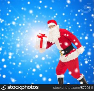 christmas, holidays and people concept - man in costume of santa claus running with gift box over blue snowy background