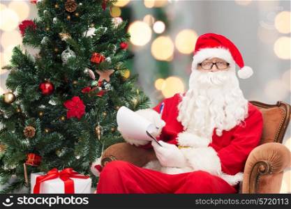 christmas, holidays and people concept - man in costume of santa claus with notepad, pen and christmas tree sitting in armchair over lights background