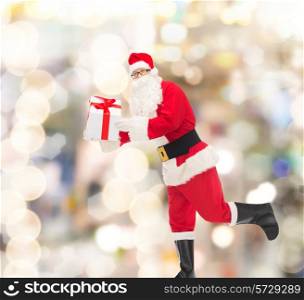 christmas, holidays and people concept - man in costume of santa claus running with gift box over lights background
