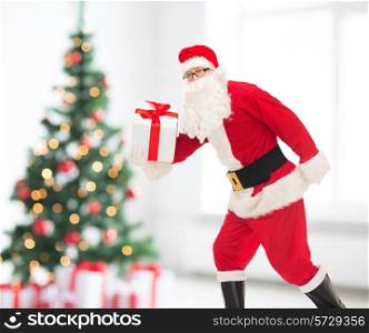 christmas, holidays and people concept - man in costume of santa claus running with gift box over living room and tree background
