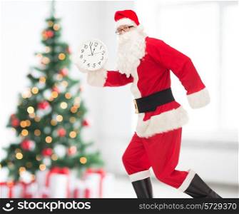 christmas, holidays and people concept - man in costume of santa claus running with clock showing twelve over living room and tree background