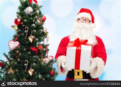 christmas, holidays and people concept - man in costume of santa claus with gift box and tree over blue lights background