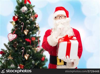christmas, holidays and people concept - man in costume of santa claus with gift box and tree making hush gesture over blue lights background