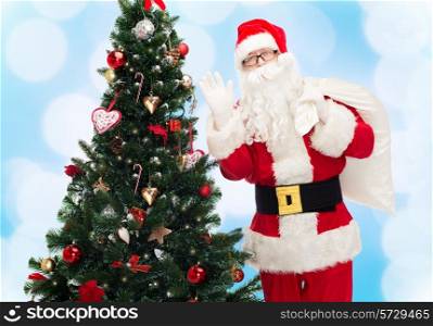 christmas, holidays and people concept - man in costume of santa claus with bag and christmas tree waving hand over blue lights background
