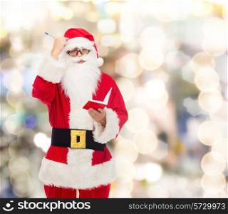 christmas, holidays and people concept - man in costume of santa claus with notepad and pen over lights background