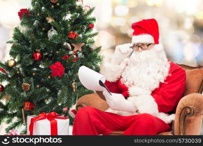 christmas, holidays and people concept - man in costume of santa claus with notepad and christmas tree sitting in armchair over lights background