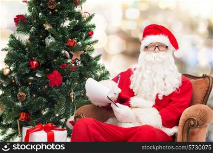 christmas, holidays and people concept - man in costume of santa claus with notepad, pen and christmas tree sitting in armchair over lights background