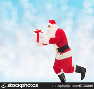 christmas, holidays and people concept - man in costume of santa claus running with gift box over blue lights background