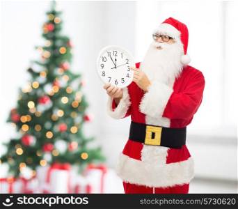 christmas, holidays and people concept - man in costume of santa claus with clock showing twelve pointing finger over living room and tree background