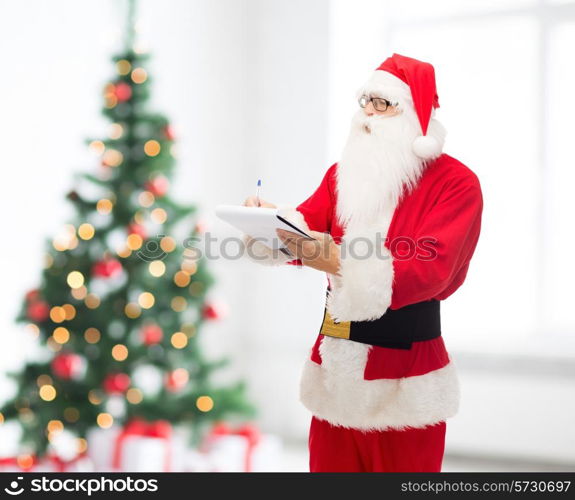 christmas, holidays and people concept - man in costume of santa claus with notepad and pen over living room and tree background