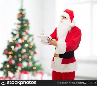 christmas, holidays and people concept - man in costume of santa claus with notepad and pen over living room and tree background