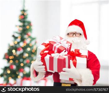 christmas, holidays and people concept - man in costume of santa claus with gift boxes over living room and tree background
