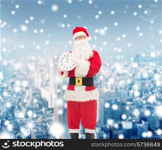 christmas, holidays and people concept - man in costume of santa claus with clock showing twelve pointing finger over snowy city background