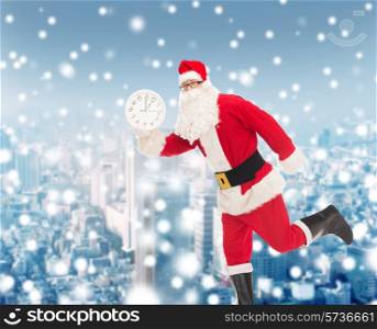 christmas, holidays and people concept - man in costume of santa claus running with clock showing twelve over snowy city background