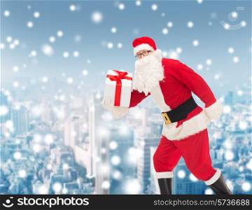 christmas, holidays and people concept - man in costume of santa claus running with gift box over snowy city background