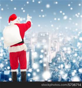 christmas, holidays and people concept - man in costume of santa claus with bag pointing finger from back over yellow lights background over snowy city background