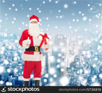 christmas, holidays and people concept - man in costume of santa claus with gift box showing thumbs up gesture over snowy city background