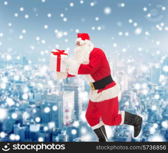 christmas, holidays and people concept - man in costume of santa claus running with gift box over snowy city background