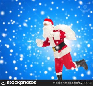 christmas, holidays and people concept - man in costume of santa claus running with bag over blue snowy background