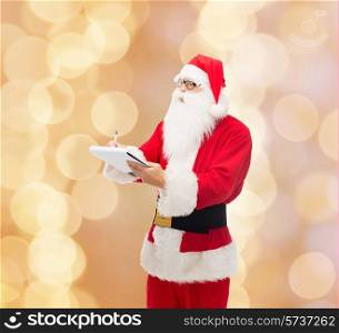 christmas, holidays and people concept - man in costume of santa claus with notepad and pen over beige lights background