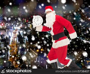 christmas, holidays and people concept - man in costume of santa claus running with clock showing twelve over snowy night city background