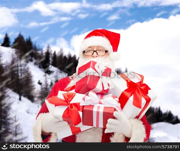 christmas, holidays and people concept - man in costume of santa claus with gift boxes over snowy mountains background