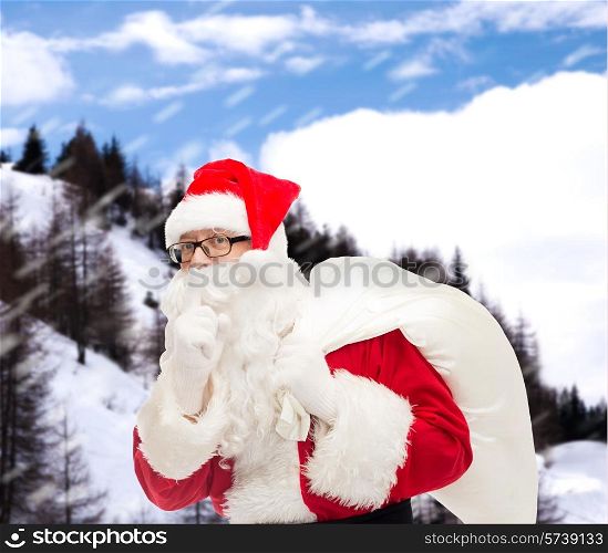 christmas, holidays and people concept - man in costume of santa claus with bag making hush gesture over snowy mountains background