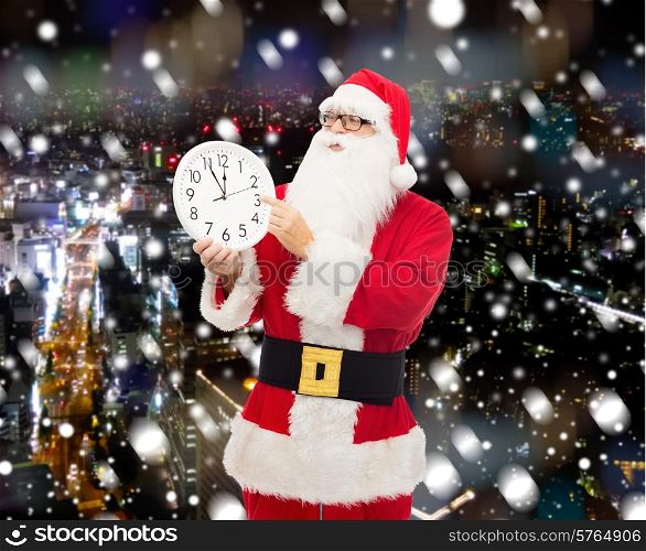 christmas, holidays and people concept - man in costume of santa claus with clock showing twelve pointing finger over snowy night city background