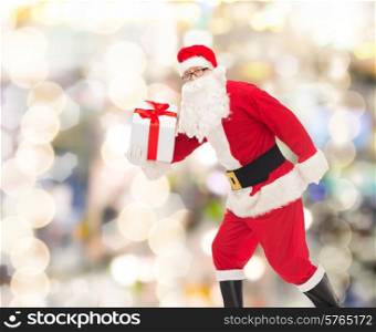 christmas, holidays and people concept - man in costume of santa claus running with gift box over lights background