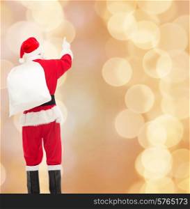 christmas, holidays and people concept - man in costume of santa claus with bag pointing finger from back over yellow lights background over beige lights background