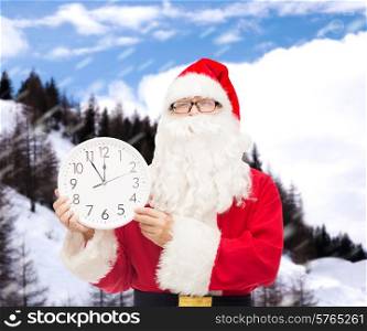 christmas, holidays and people concept - man in costume of santa claus with clock showing twelve over snowy mountains background
