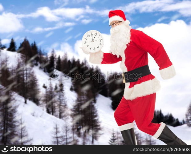 christmas, holidays and people concept - man in costume of santa claus running with clock showing twelve over snowy mountains background