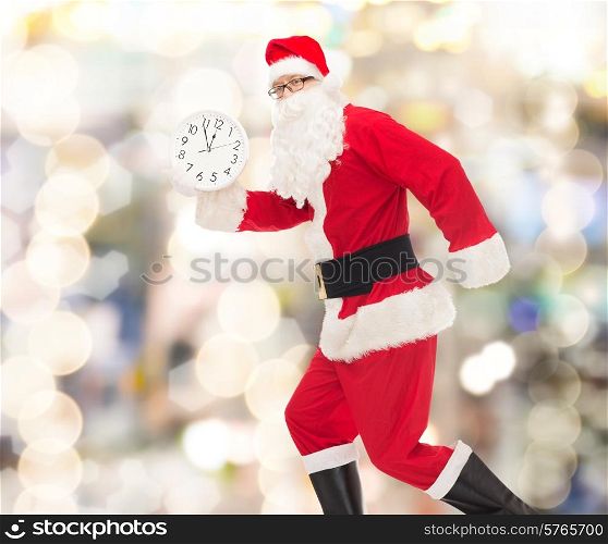 christmas, holidays and people concept - man in costume of santa claus running with clock showing twelve over lights background