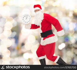 christmas, holidays and people concept - man in costume of santa claus running with clock showing twelve over lights background