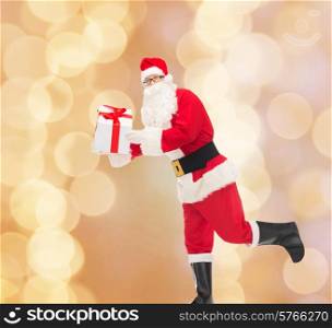 christmas, holidays and people concept - man in costume of santa claus running with gift box over beige lights background