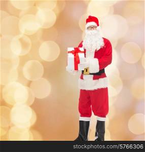 christmas, holidays and people concept - man in costume of santa claus with gift box over beige lights background