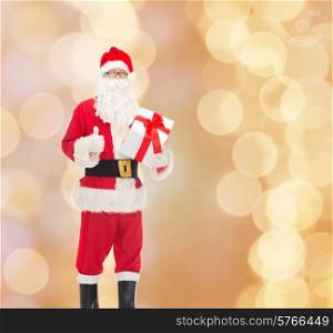 christmas, holidays and people concept - man in costume of santa claus with gift box showing thumbs up gesture over beige lights background