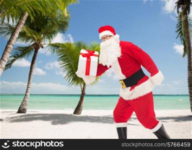 christmas, holidays and people concept - man in costume of santa claus running with gift box over tropical beach background