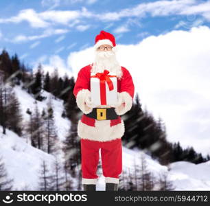 christmas, holidays and people concept - man in costume of santa claus with gift box over snowy mountains background