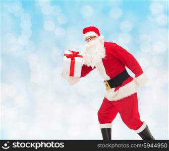 christmas, holidays and people concept - man in costume of santa claus running with gift box over blue lights background