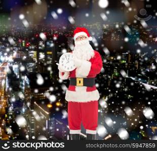 christmas, holidays and people concept - man in costume of santa claus with clock showing twelve pointing finger over snowy night city background