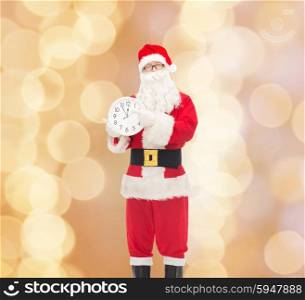 christmas, holidays and people concept - man in costume of santa claus with clock showing twelve pointing finger over beige lights background