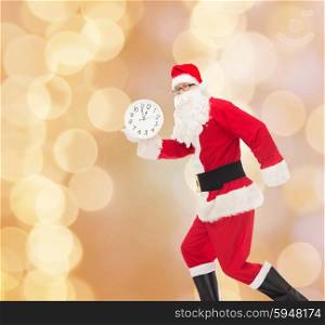 christmas, holidays and people concept - man in costume of santa claus running with clock showing twelve over beige lights background