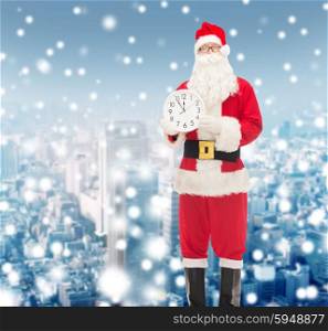 christmas, holidays and people concept - man in costume of santa claus with clock showing twelve over snowy city background