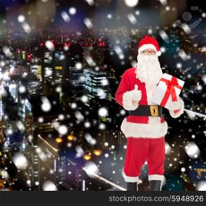 christmas, holidays and people concept - man in costume of santa claus with gift box showing thumbs up gesture over snowy night city background
